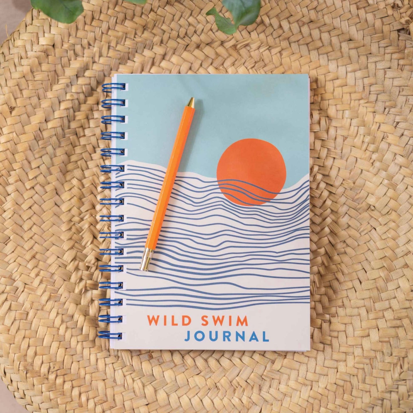 A spiral-bound swim journal with a blue, orange, and white cover design, featuring a pen and placed on a woven mat. The journal is labeled 'Wild Swim Journal'.