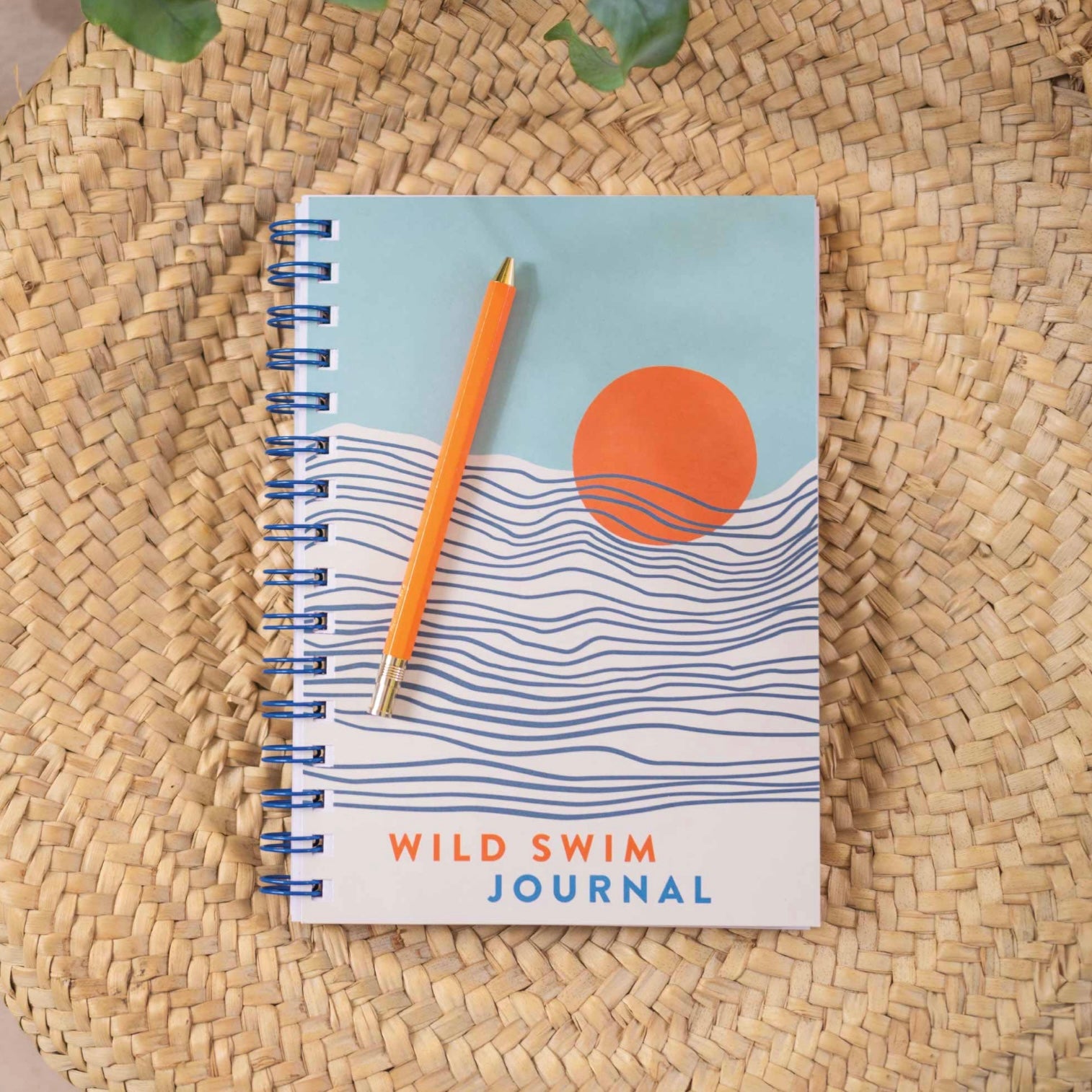A spiral-bound swim journal with a blue, orange, and white cover design, featuring a pen and placed on a woven mat. The journal is labeled 'Wild Swim Journal'.