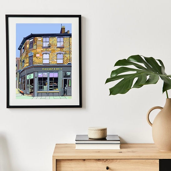 A framed A4 print depicting a colorful building, hanging on a white wall above a shelf with books and a plant vase.